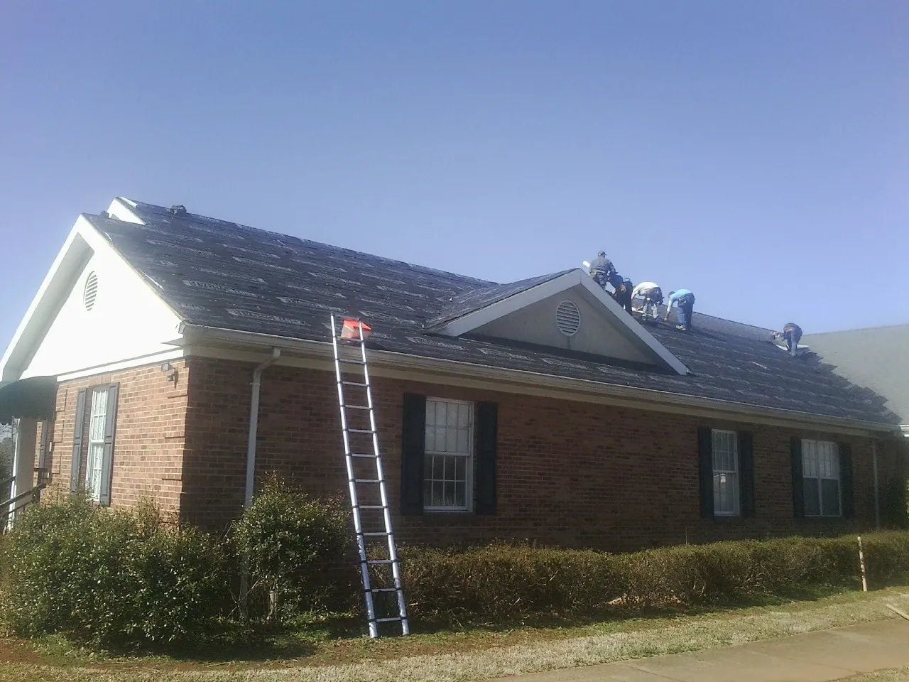 Crew tearing shingles off a brick ranch roof with a ladder set — Emergency Roof Repair work by our crew in Dewitt