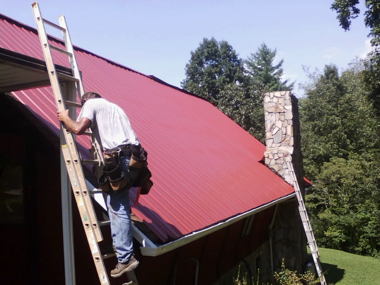 Roofer on a ladder beside a red metal roof and stone chimney — Soffit & Fascia Repair by Roofing Brothers in Dewitt